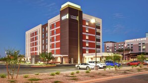 A multi-story hotel with a prominent 'HOME' sign, a parking lot with several cars, and a 'Hyatt Place' building in the background. - Home2 Suites by Hilton Gilbert (Gilbert)