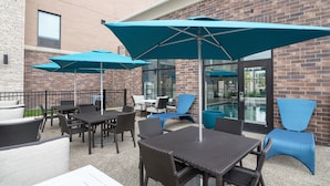 An outdoor patio with tables and chairs, blue umbrellas, and a brick building in the background. - Hampton Inn & Suites Allen Park (Allen Park)
