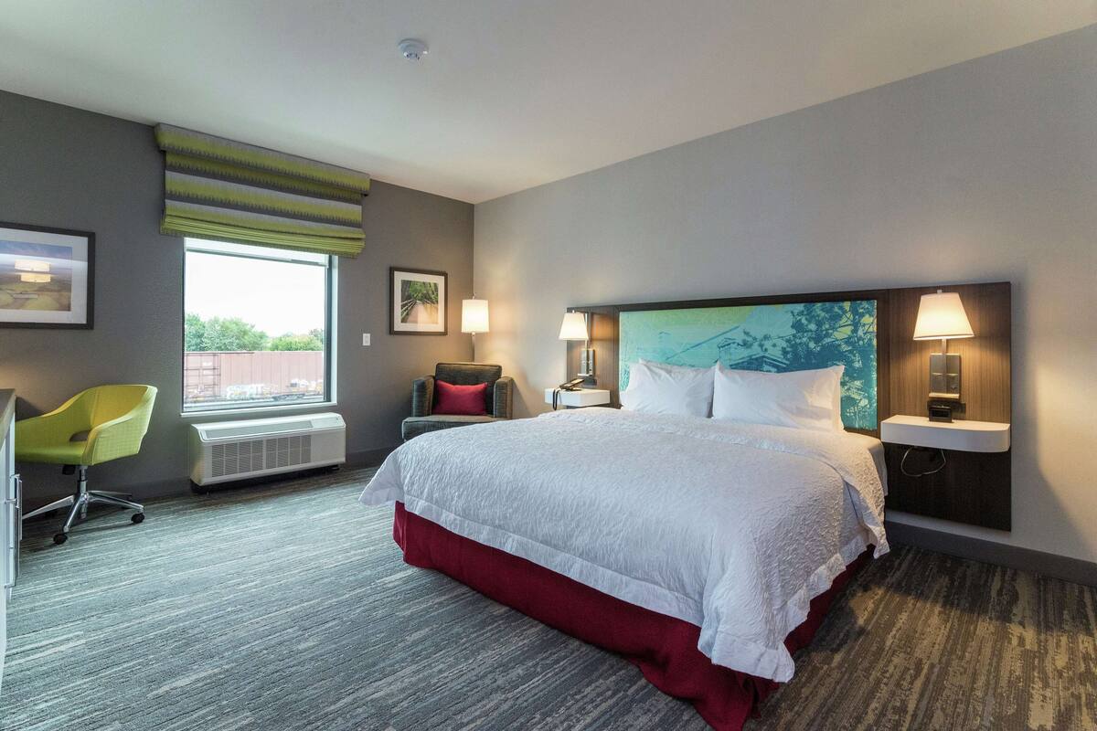 Une chambre d’hôtel avec un grand lit, un bureau avec une chaise, une fenêtre donnant sur un beau paysage et une lampe fixée au mur.