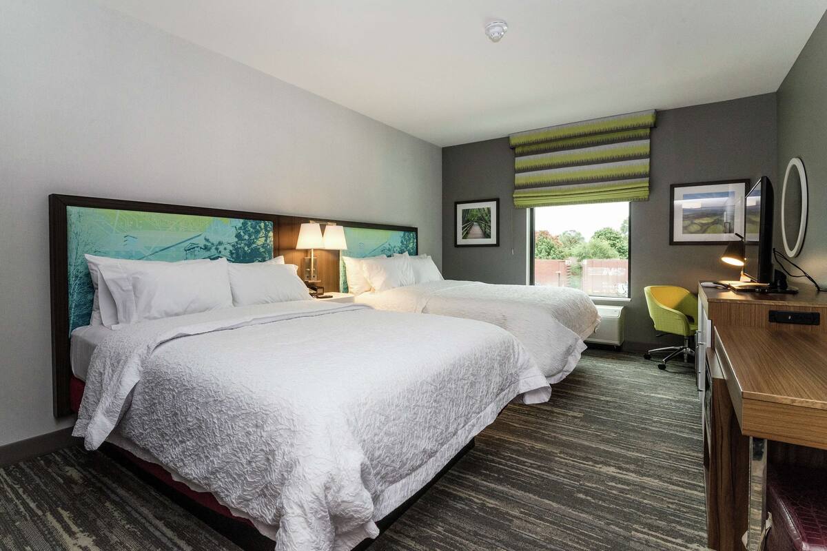 Une chambre d’hôtel avec deux lits, un bureau, une chaise, une fenêtre donnant sur un beau paysage et un tableau au mur.