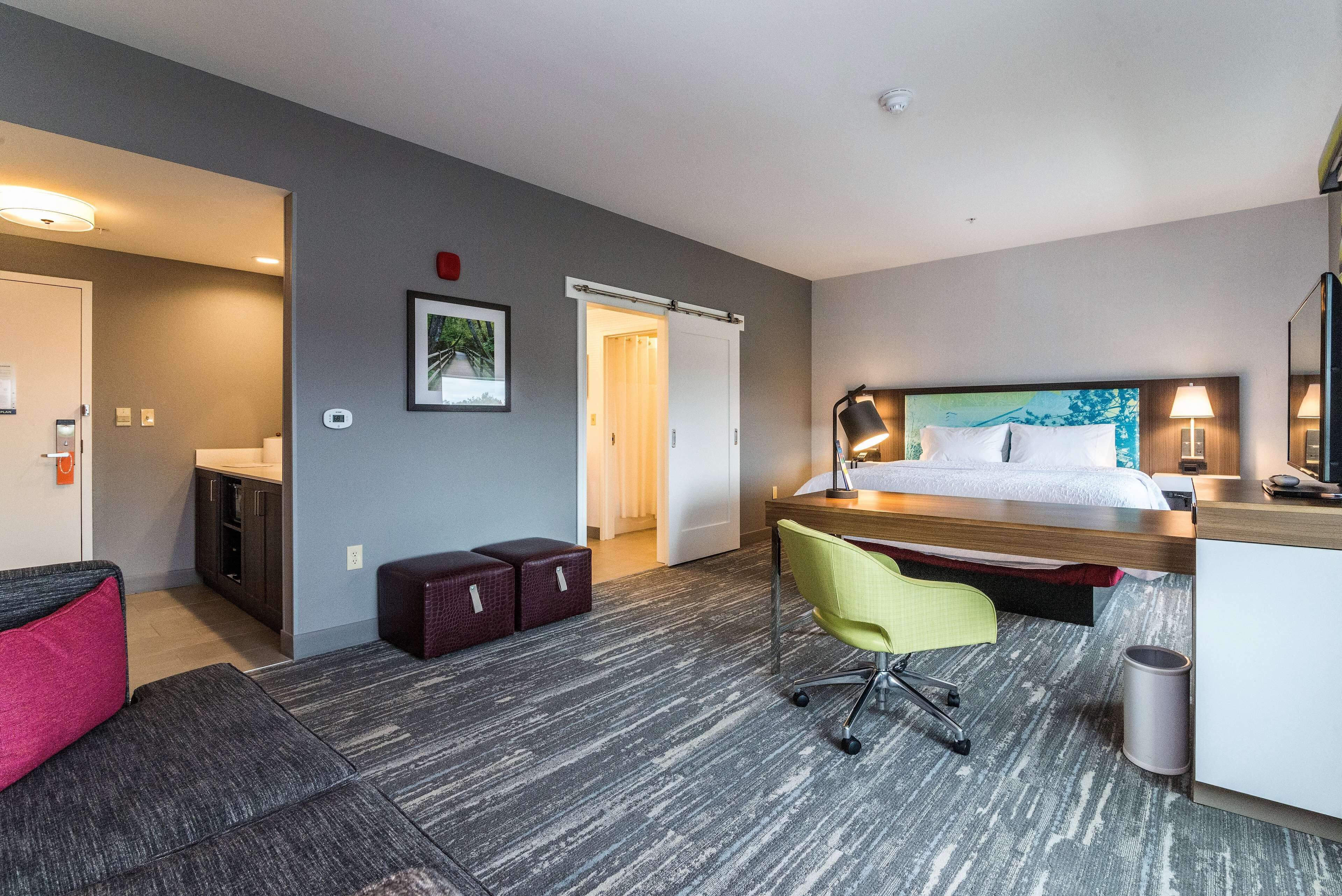 A hotel room with a bed, desk, chair, and a small kitchenette.