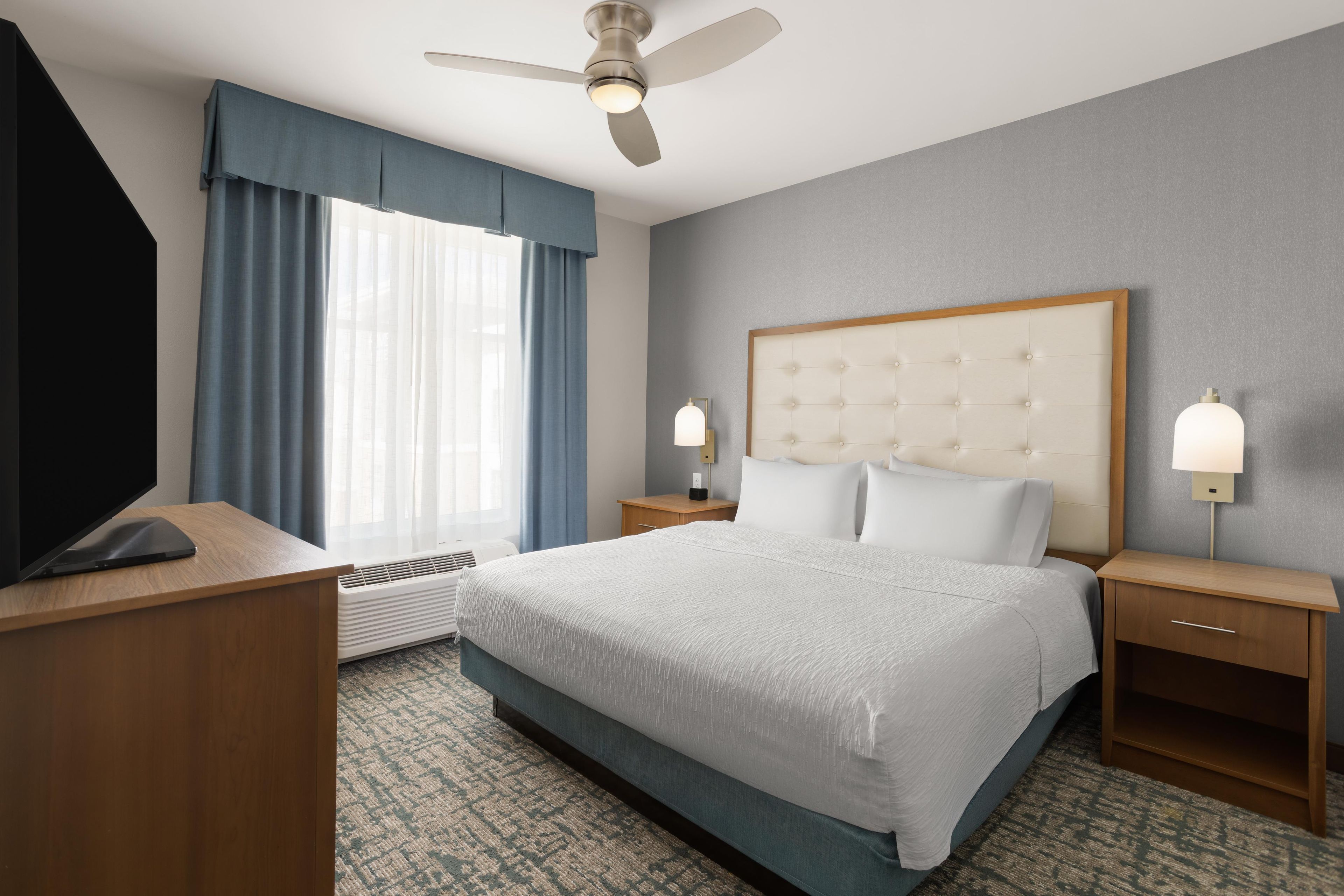 Quarto de hotel com cama, mesas de cabeceira, escrivaninha, ventoinha no teto e janela com cortinas.