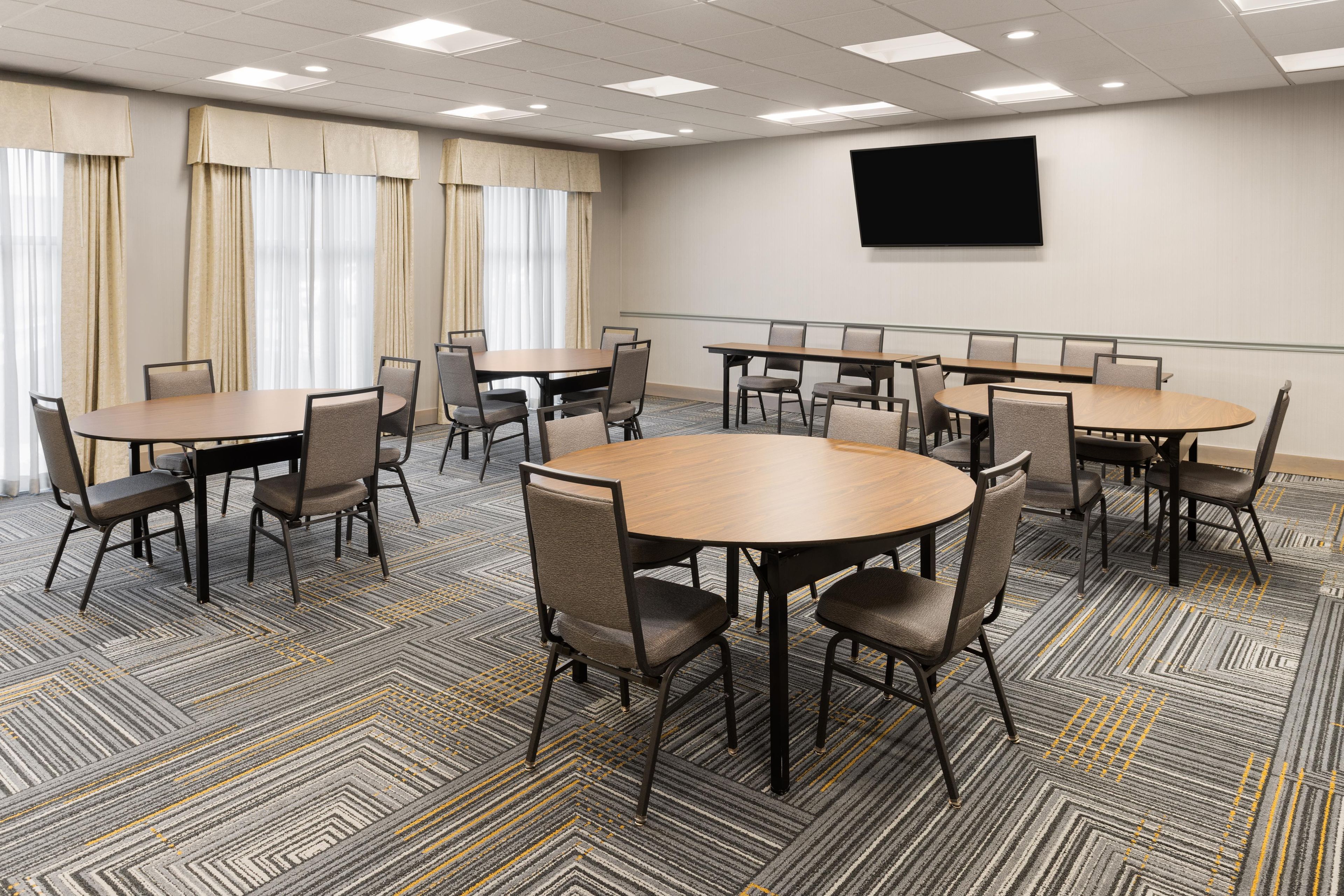 A conference room with multiple tables and chairs, a flat-screen TV on the wall, and large windows with curtains.
