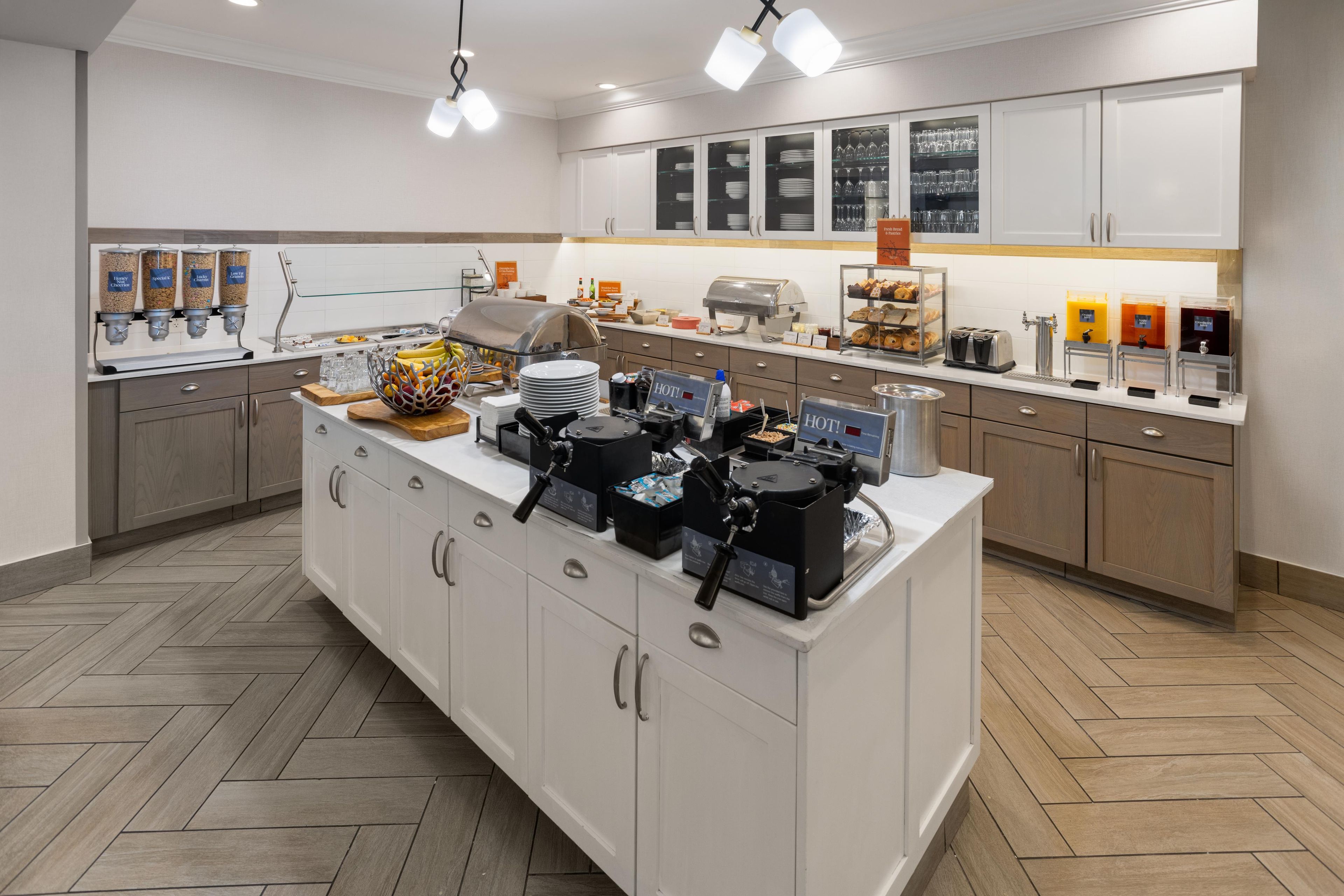 A modern kitchen with a central island, equipped with coffee machines, a food display, and a beverage station.