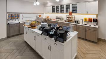 A modern kitchen with a central island, equipped with coffee machines, a food display, and a beverage station.