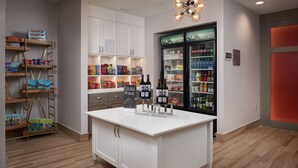 Un vestíbulo de hotel con una isla central, muebles blancos, un refrigerador para bebidas y estantes llenos de aperitivos y bebidas.