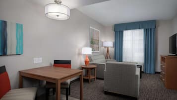 A hotel room with a wooden dining table, two chairs, a sofa, a TV stand, and a lamp.
