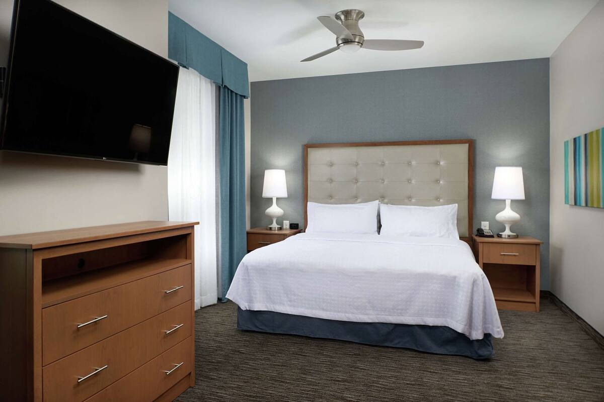 A hotel room with a bed, bedside tables with lamps, a TV mounted on the wall, a wooden dresser, and a ceiling fan.