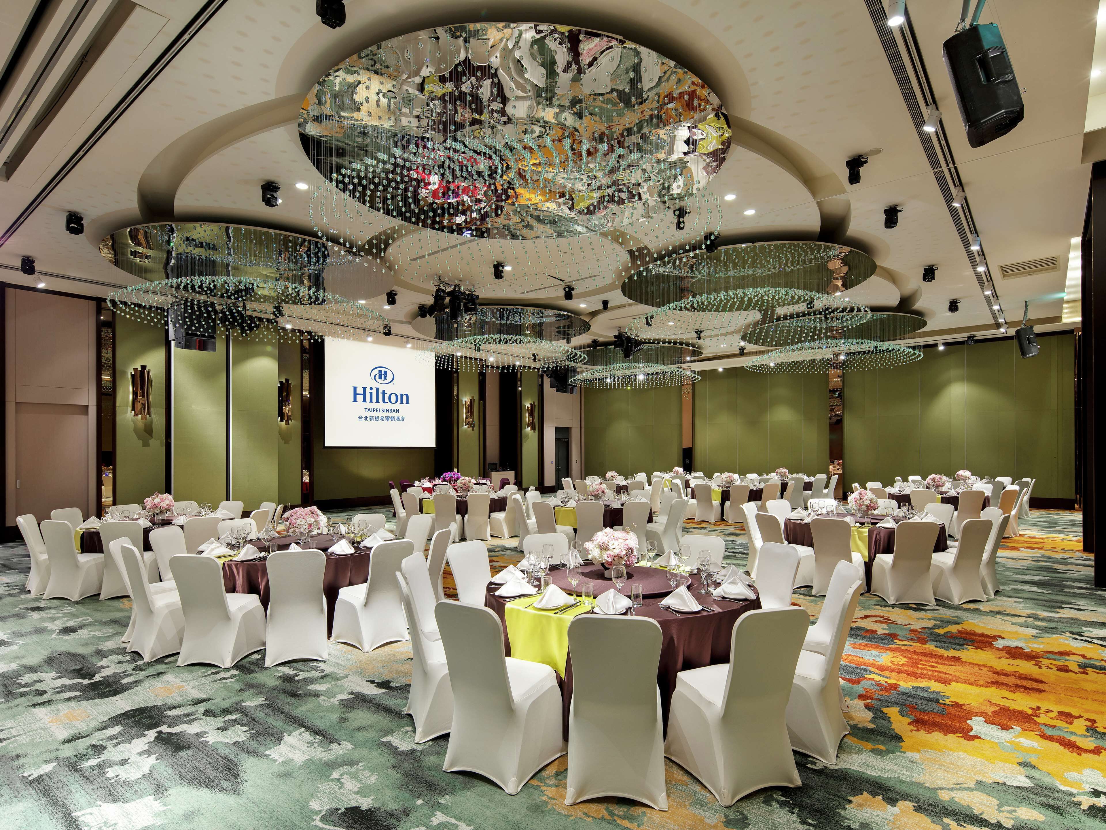 Ein Festsaal mit runden Tischen, weißen Stühlen und einer großen Leinwand, die 'Hilton Taipei Sinban' anzeigt.