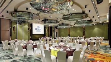 Une salle de banquet avec des tables rondes, des chaises blanches et un grand écran affichant le texte « Hilton Taipei Sinban ».