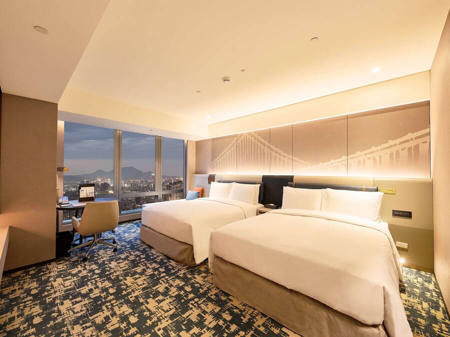 A hotel room with two beds, a desk, and a large window offering a city view.