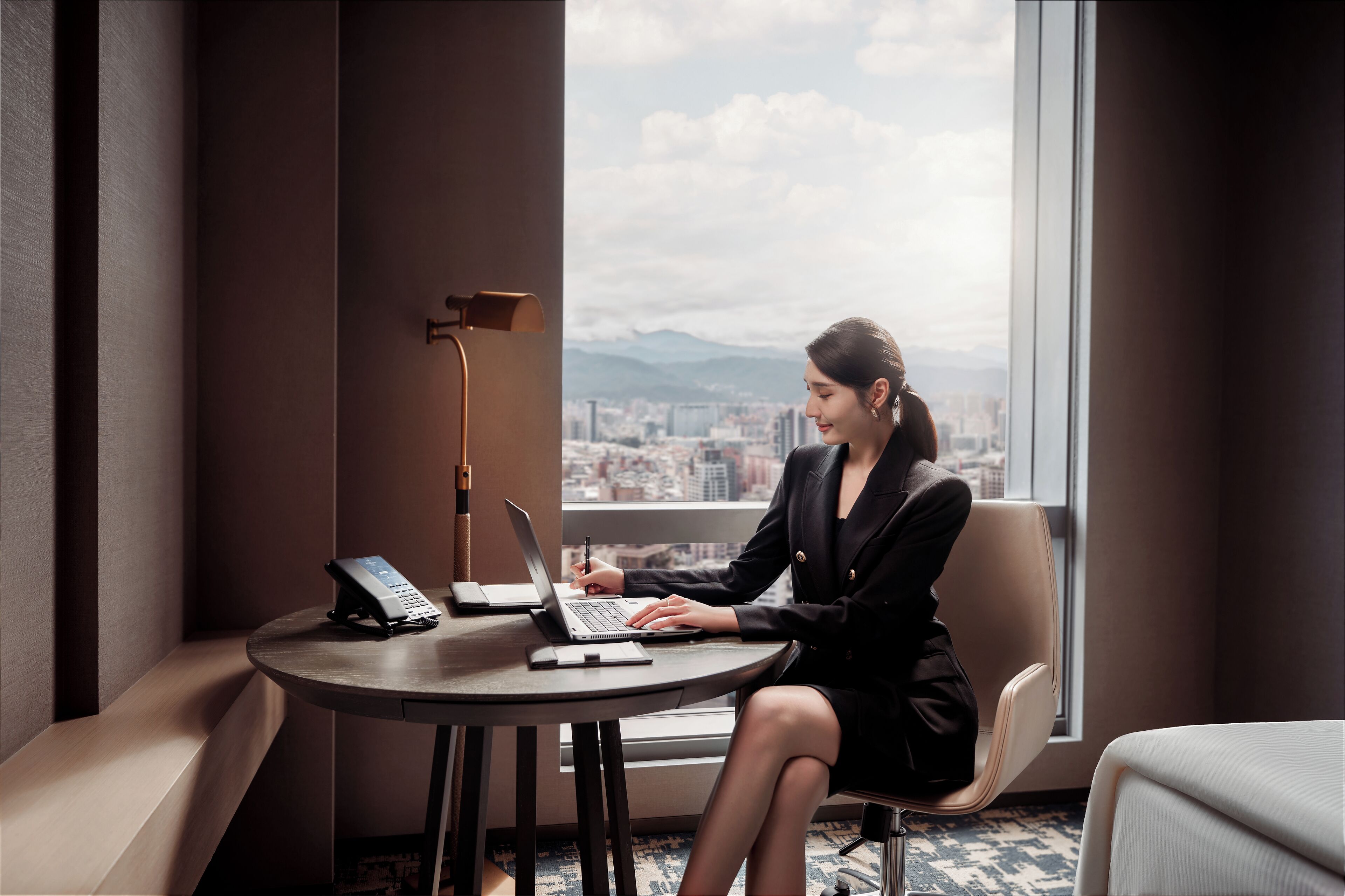 A woman sitting at a round table with a laptop and a phone, in a room with a large window overlooking a cityscape.