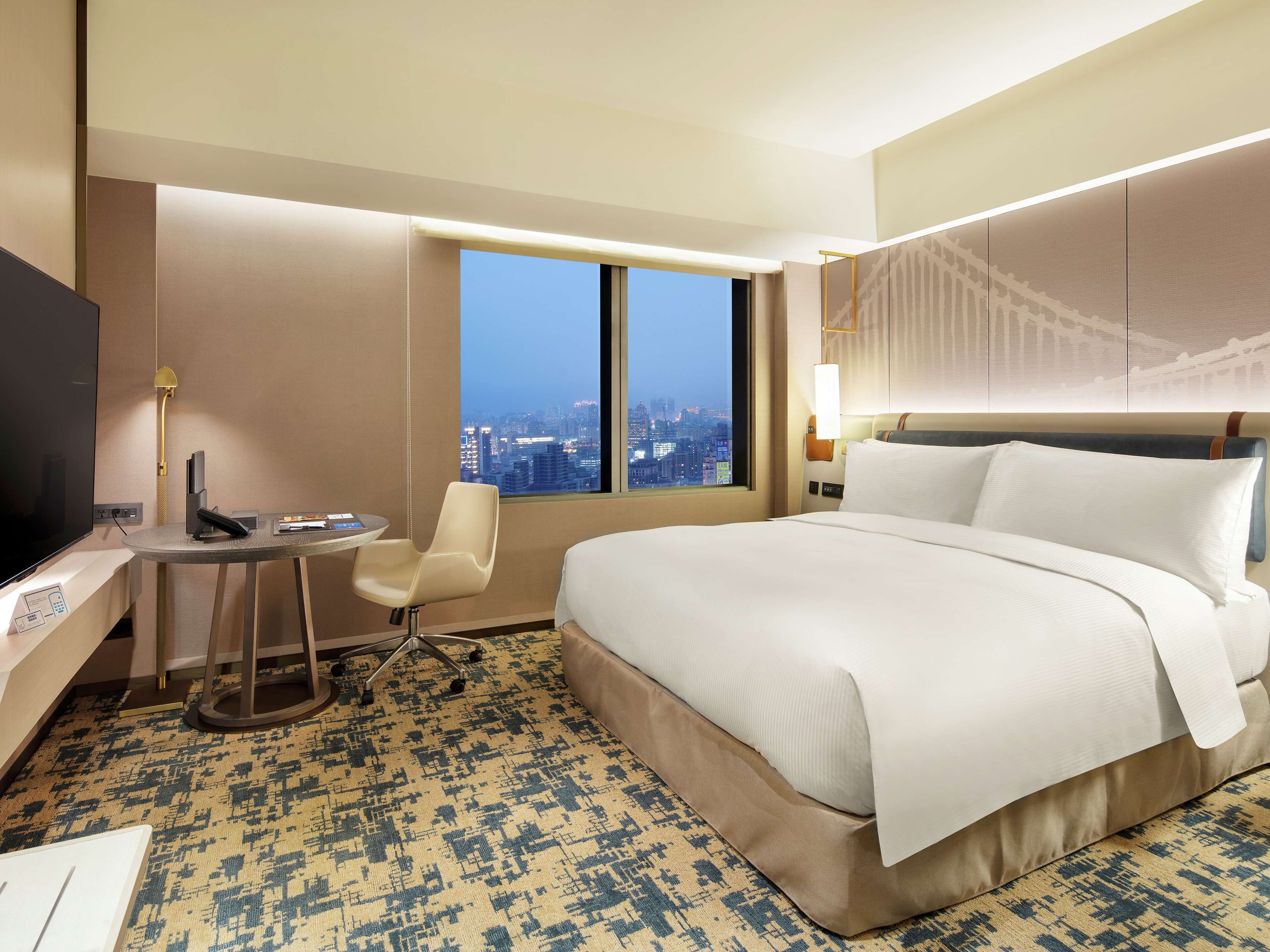 A hotel room with a large bed, a desk with a chair, a TV, and a window with a city view.