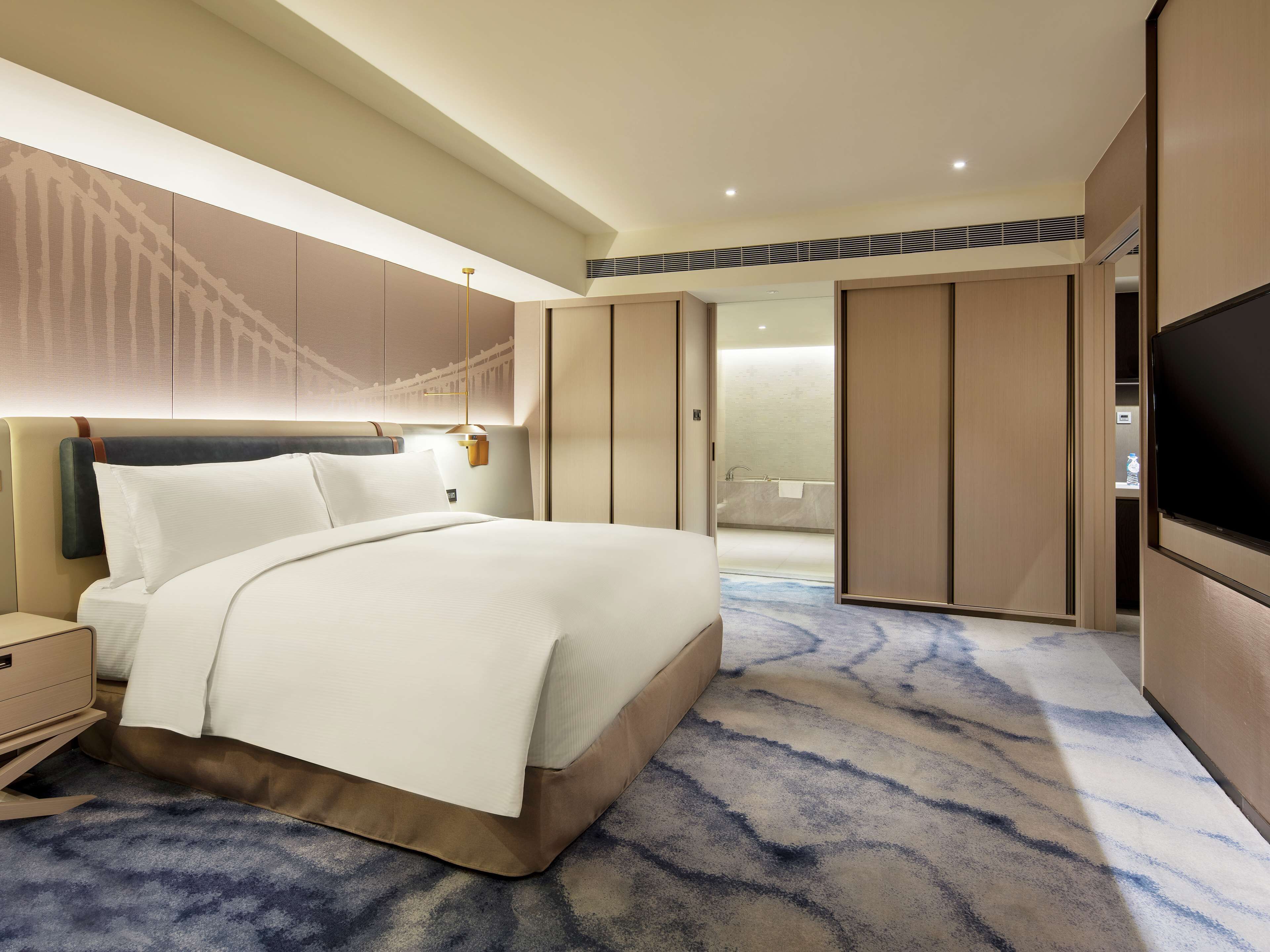 A modern hotel room with a large bed, a TV, a wardrobe, and a bathroom visible through an open door.