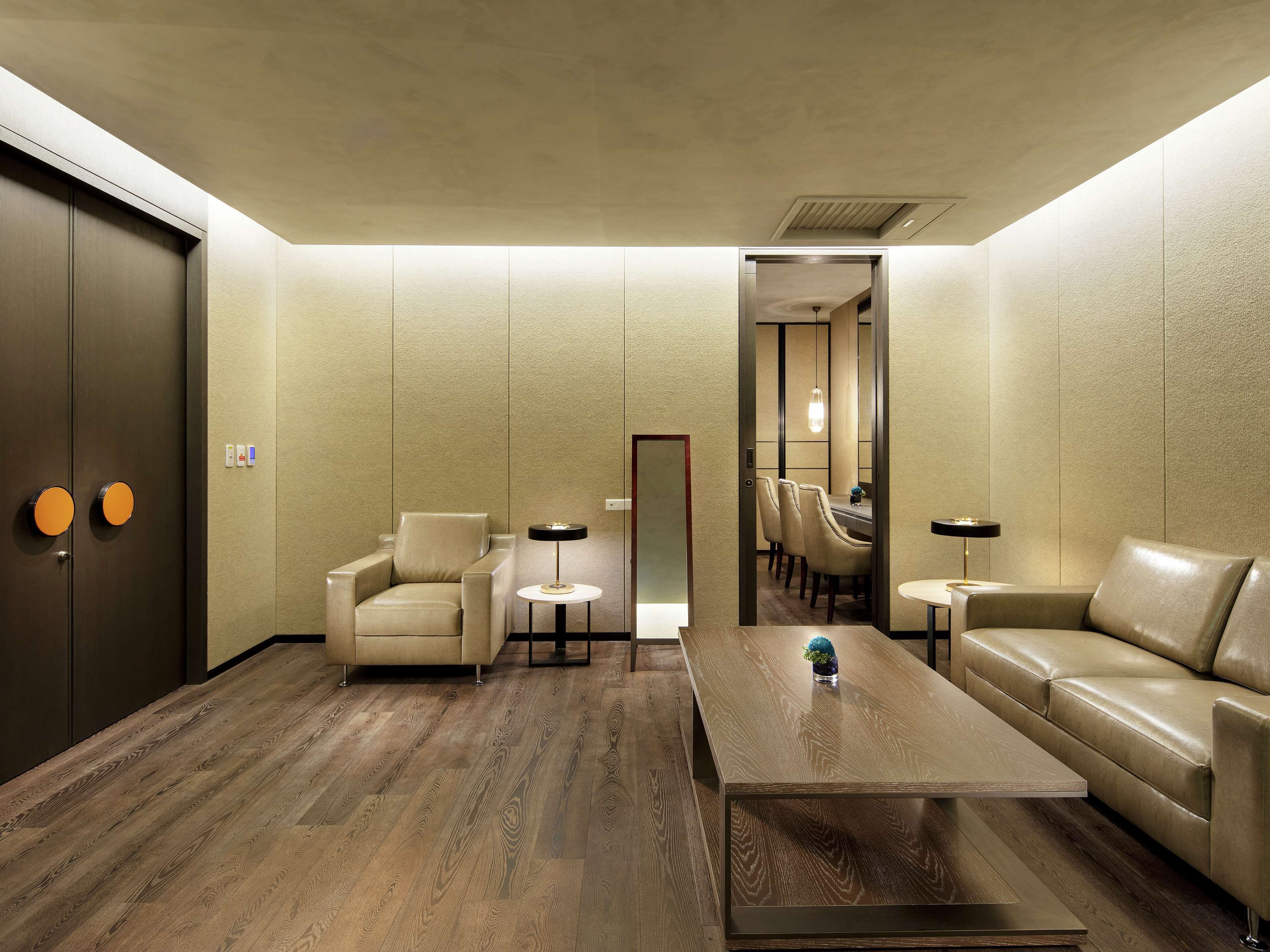 A modern hotel lobby with a wooden floor, leather seating, a coffee table, and a view into a dining area.