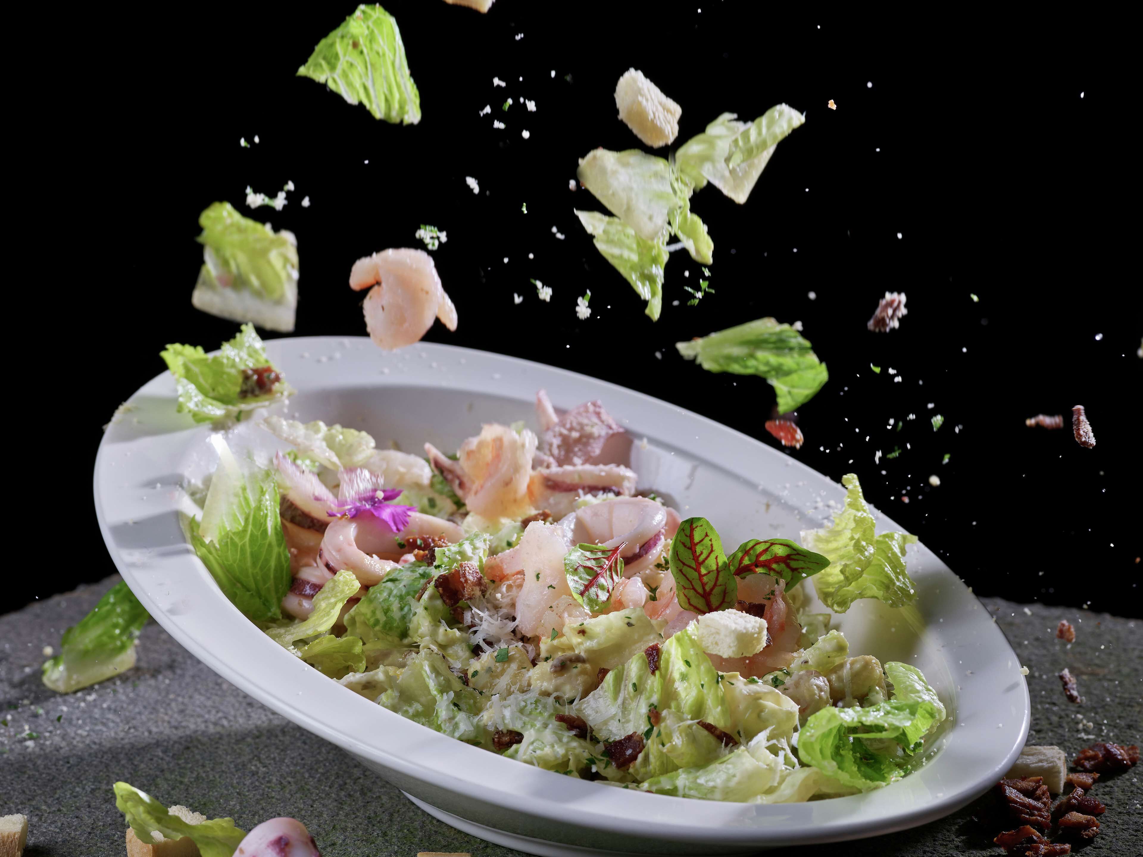 Un piatto di insalata con lattuga, gamberi e pancetta, con alcune foglie di lattuga che volano.