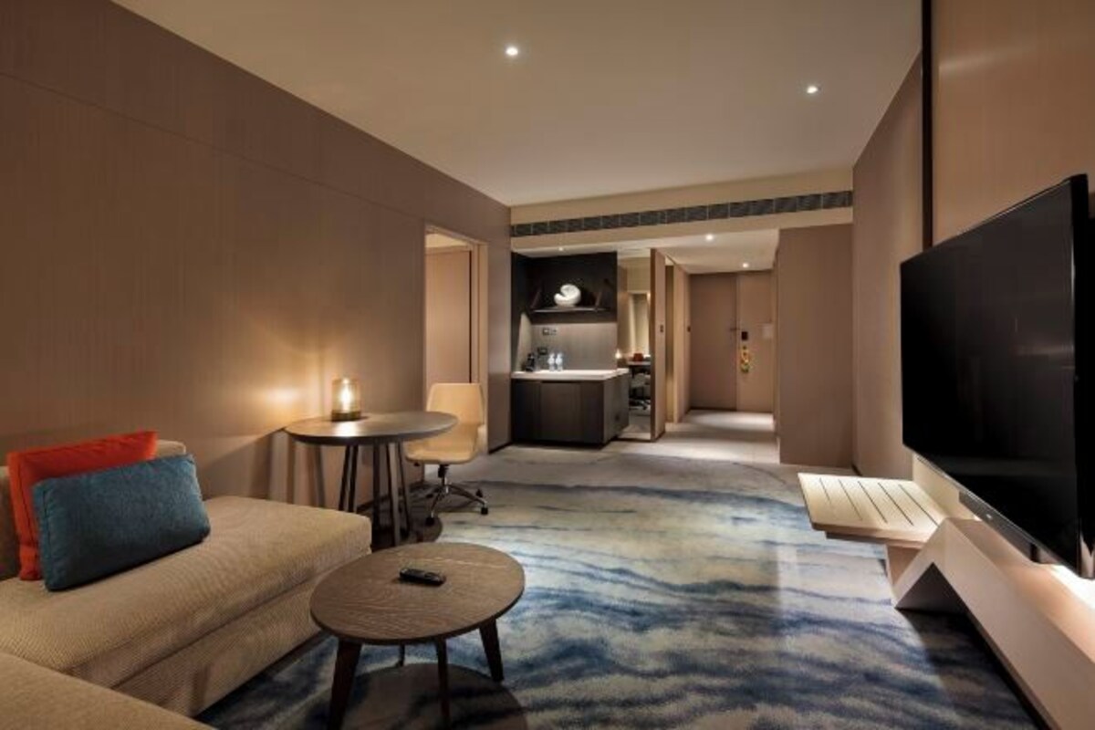 A modern hotel room with a sofa, a small round table, a TV, and a kitchen area in the background.