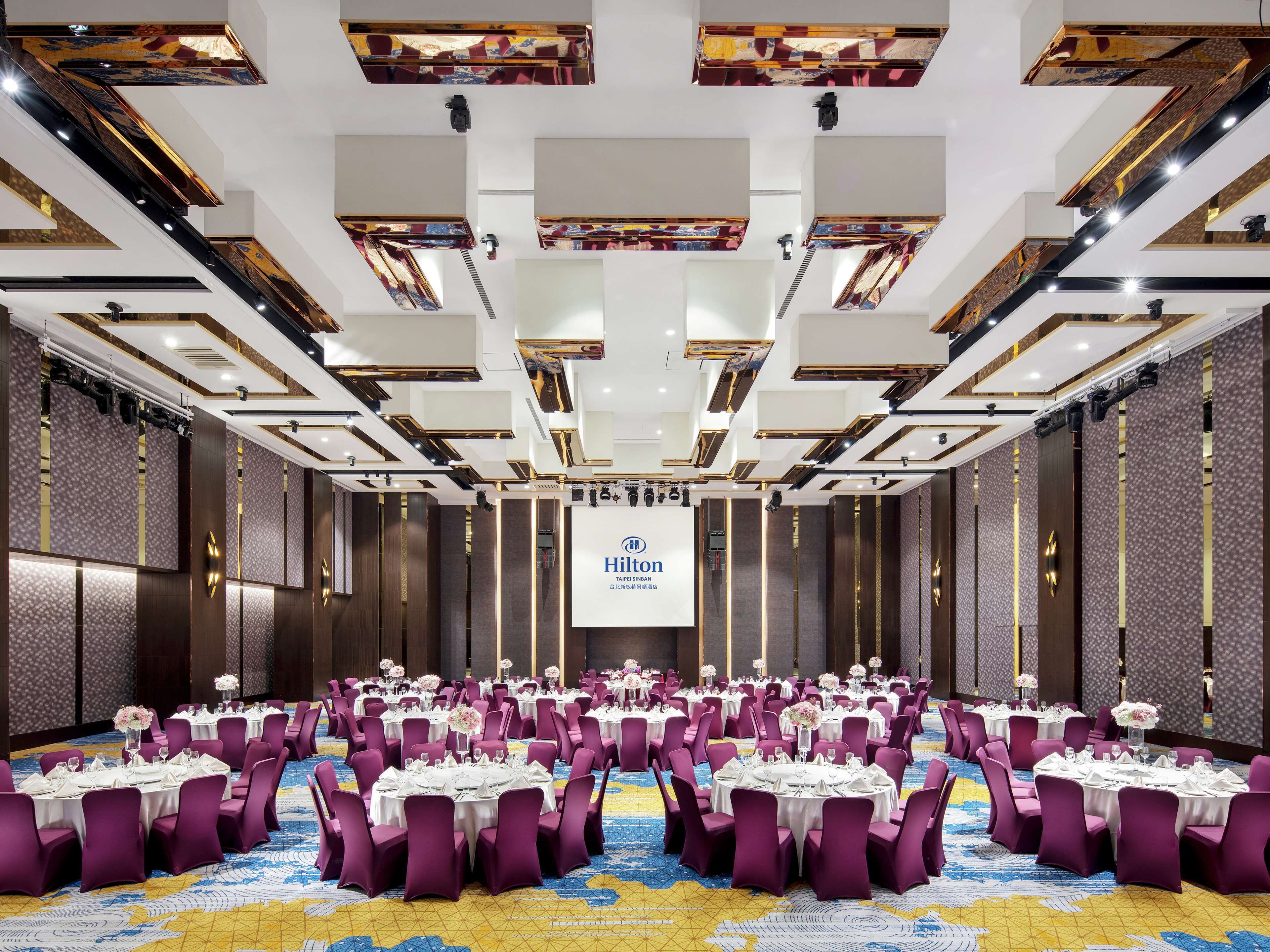 A large conference room with round tables, purple chairs, and a central screen displaying the Hilton logo.