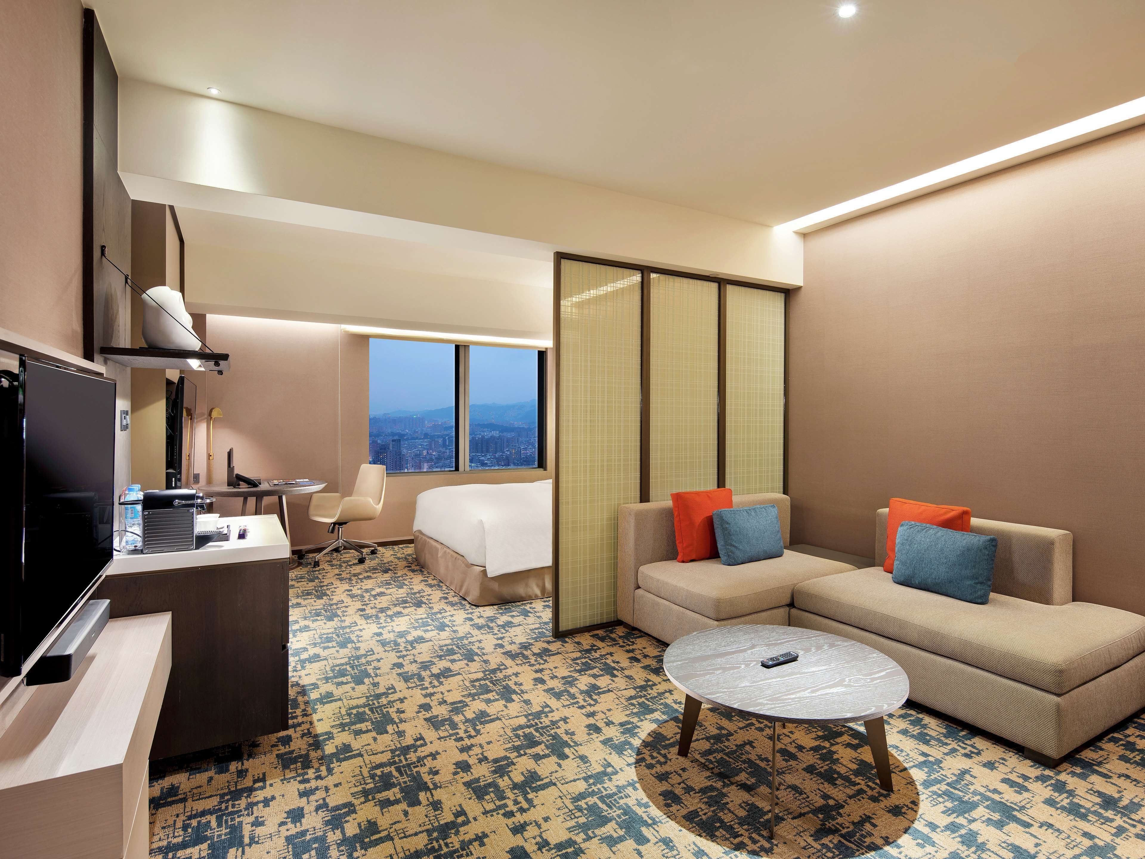 A hotel room with a bed, a sofa, a small table, a desk, and a television.