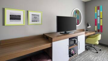 A modern office desk with a flat-screen monitor, a lamp, and a chair. There are framed pictures on the wall and a colorful abstract art piece.