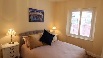 A bedroom with a bed, two bedside lamps, a nightstand, a window with blinds, and a wall-mounted picture.