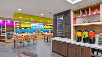 A modern hotel lobby with a kitchen area, seating, and a coffee station.