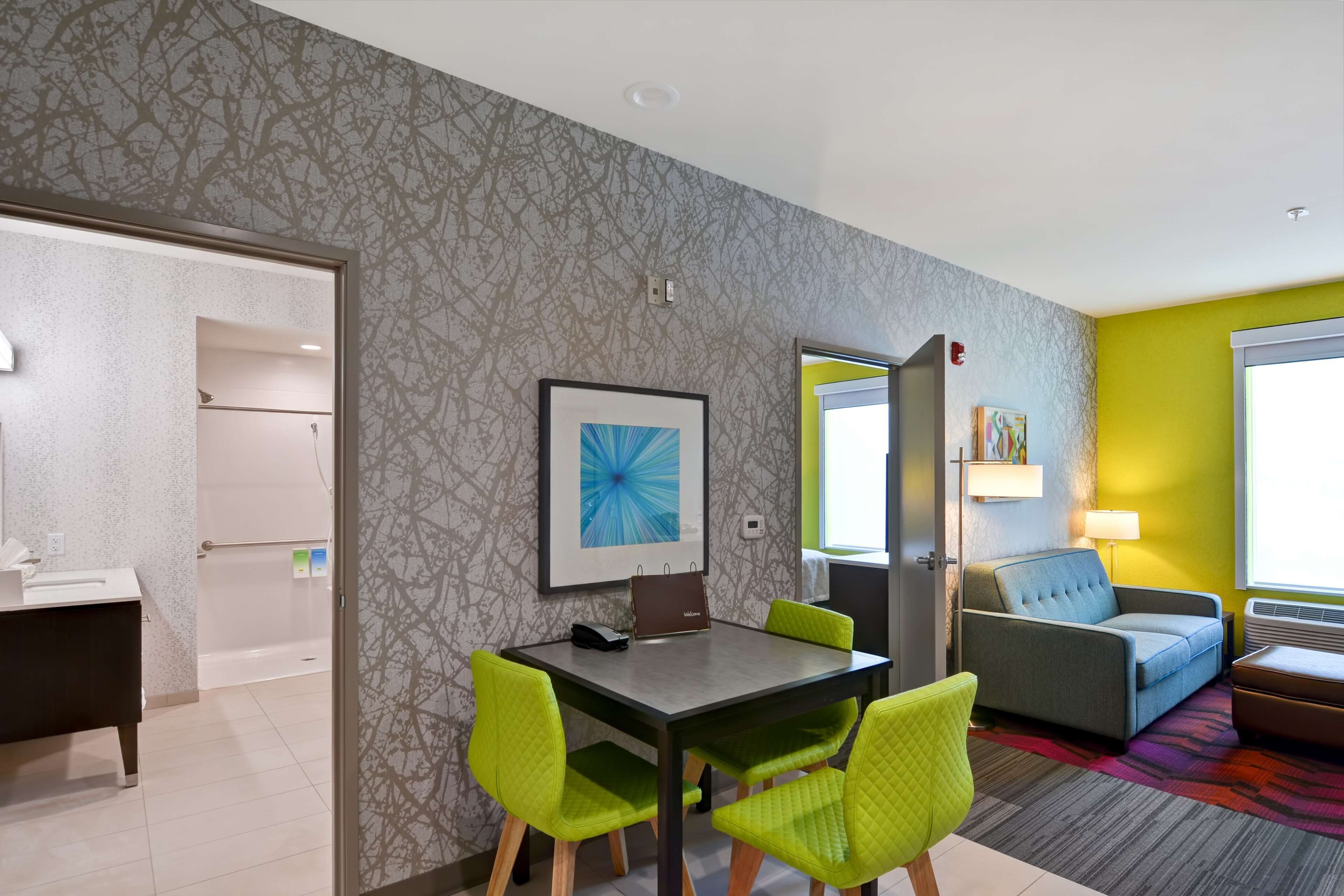 Une chambre d’hôtel avec salle de bain, un coin repas avec une table et des chaises, un canapé et un tapis coloré.