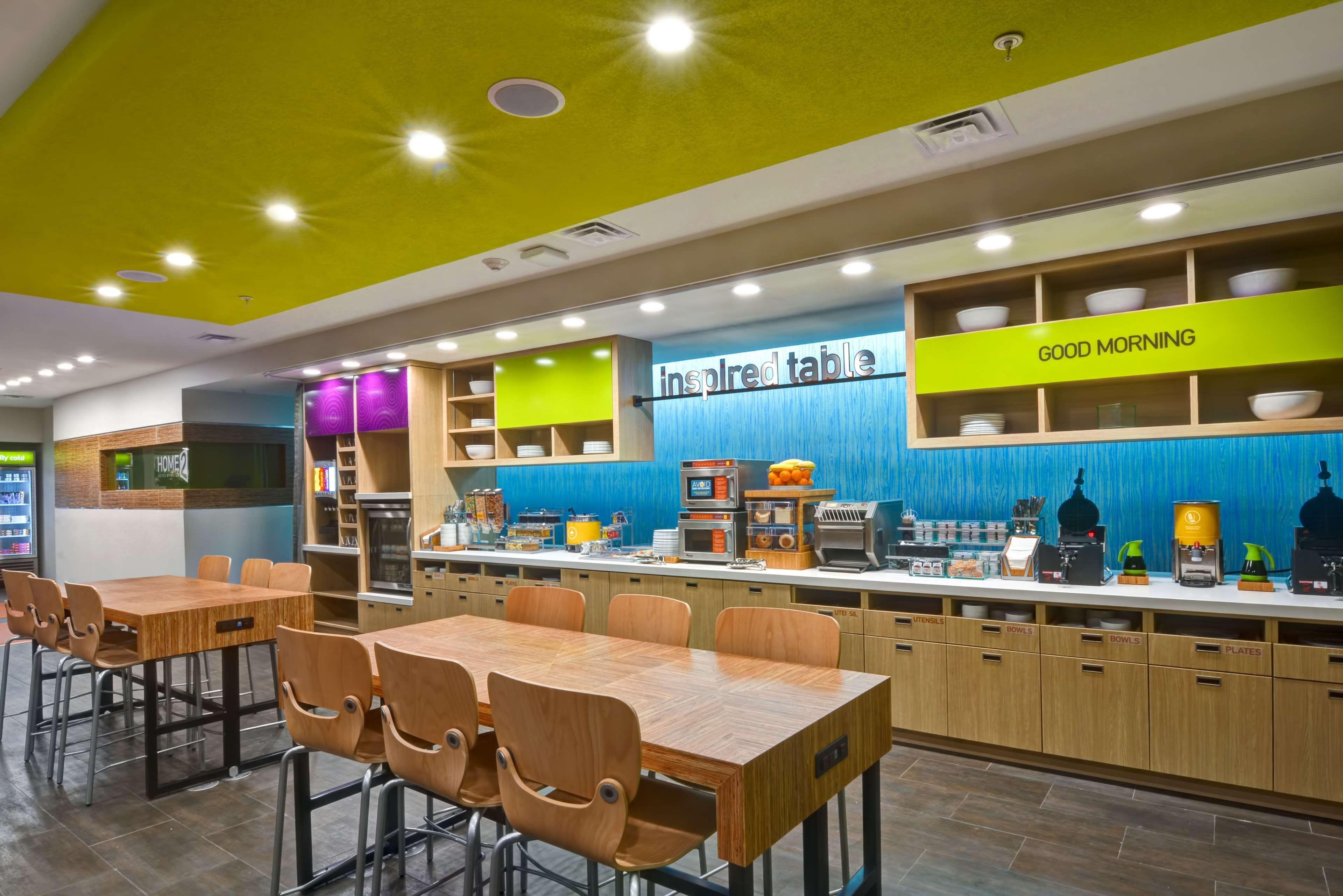 Un café moderne avec un plafond jaune vif, un mur bleu sur lequel on peut lire les mots « table d’inspiration », et un comptoir où sont exposés des appareils de cuisine et des ustensiles.