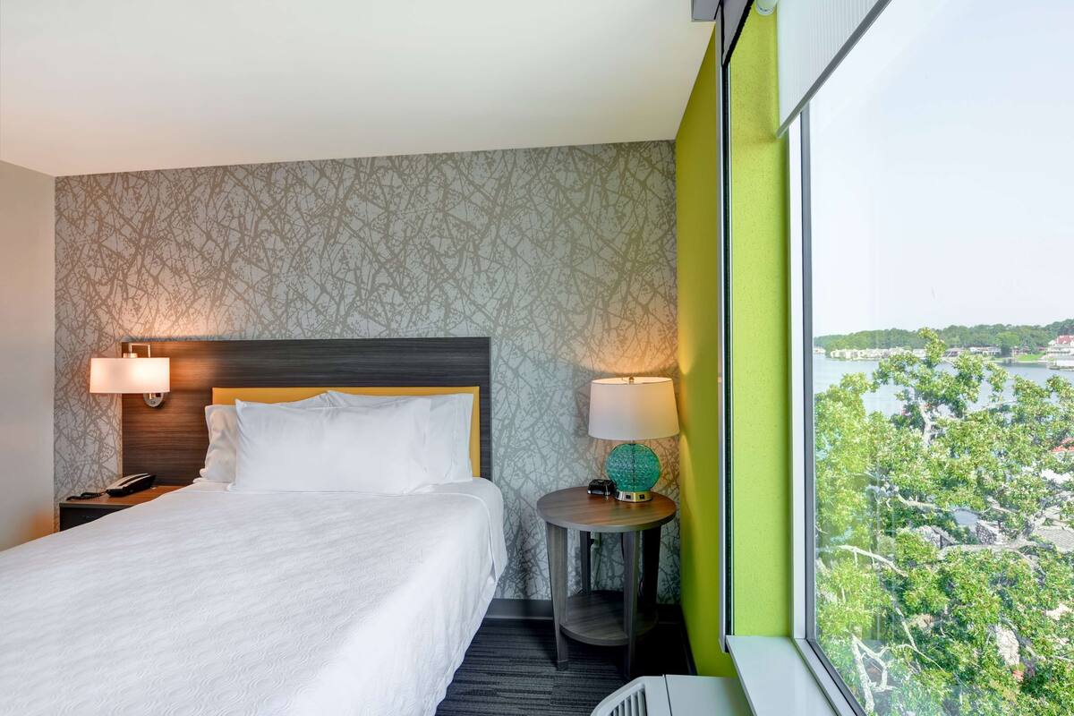 A hotel room with a large bed, two bedside lamps, a round table with a lamp, and a view of a cityscape through a large window.