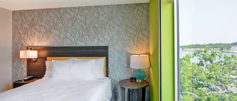 Ein Hotelzimmer mit einem großen Bett, zwei Nachttischlampen, einem runden Tisch inklusive Lampe und Blick auf eine Stadtlandschaft durch ein großes Fenster.