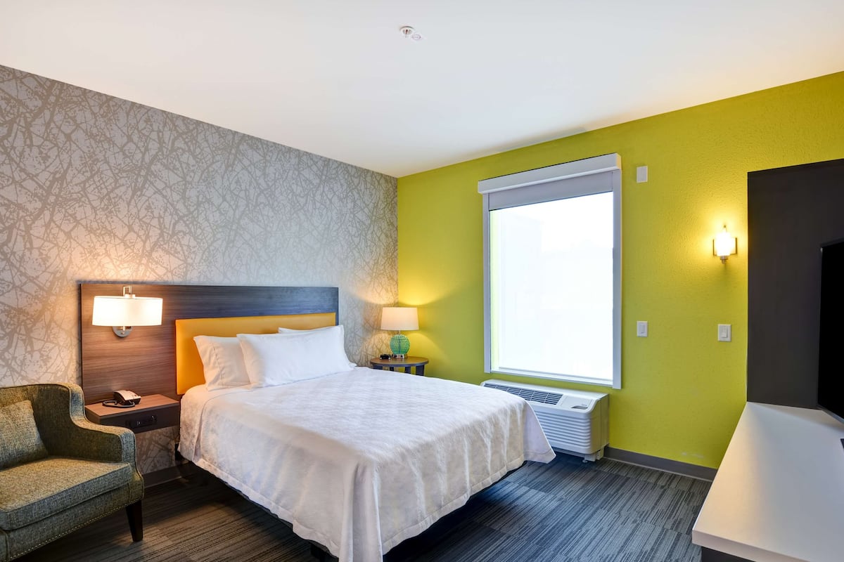A hotel room with a bed, nightstand, lamp, chair, and a large window.