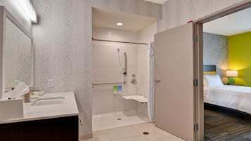 A hotel bathroom with a shower, a sink, and a toilet. There is a bed visible through an open door.