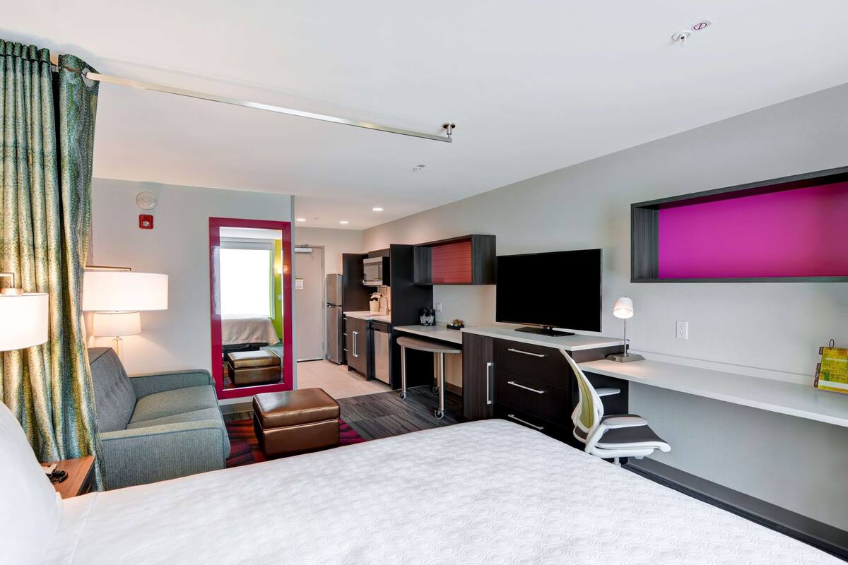 A modern hotel room with a bed, a sofa, a desk, a TV, and a kitchenette.