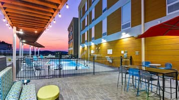 Zona de piscina al aire libre con asientos y mesas, rodeada por una cerca, y un edificio con paredes y ventanas amarillas.