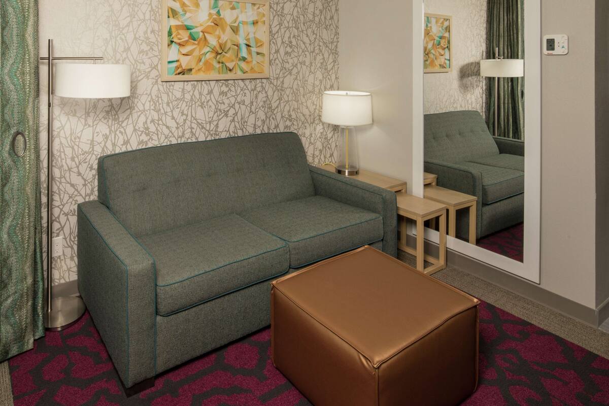 A hotel room with a grey sofa, a brown ottoman, a small table, two lamps, and a mirror reflecting the room.
