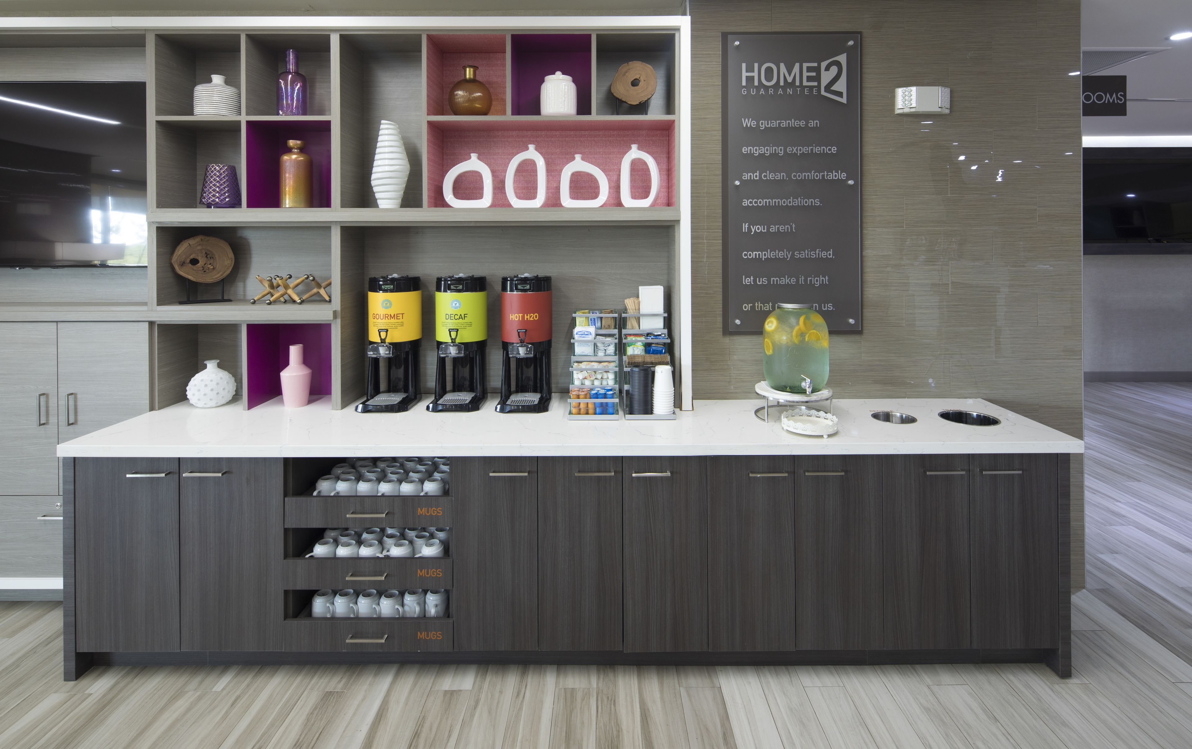 Una moderna zona de recepción de hotel con mostrador, máquinas de café y un cartel que indica una garantía de hogar.