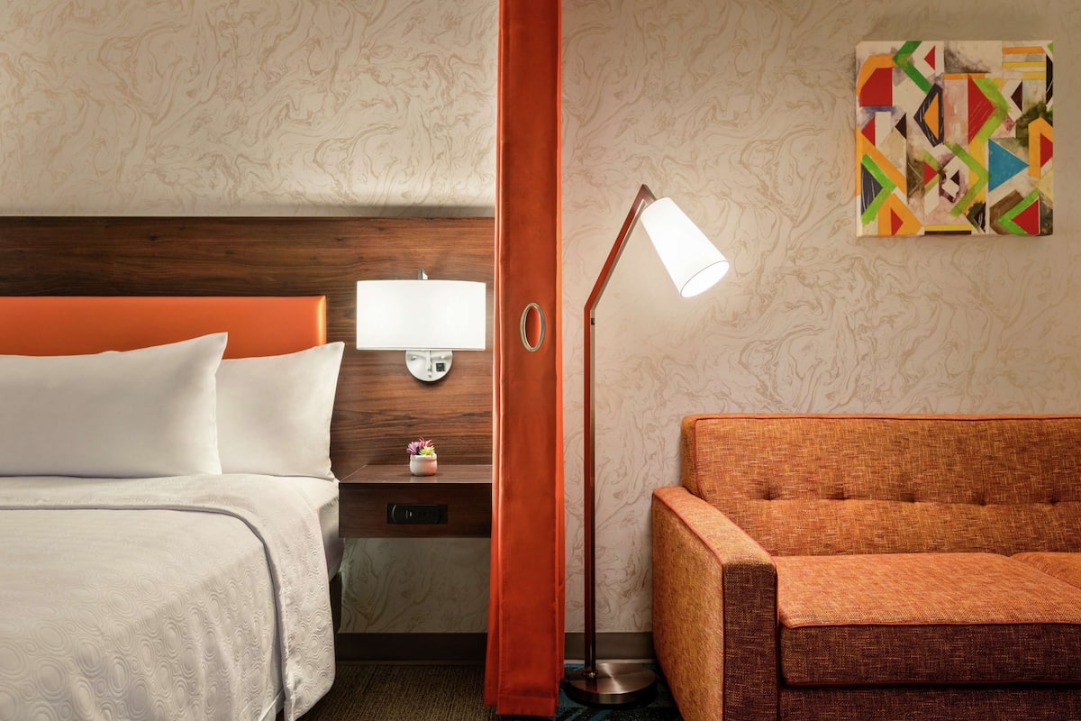 A hotel room with a bed, a nightstand, a lamp, a sofa, and a decorative wall piece.