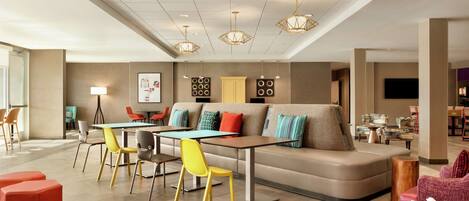 A modern lounge area with a sofa, tables, and chairs, featuring a flat-screen TV and a coffee machine.