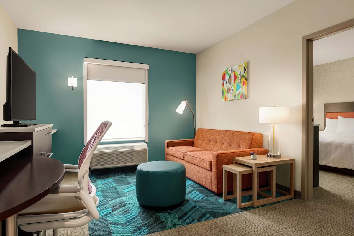 A hotel room with a teal accent wall, a sofa, a small ottoman, a desk with a chair, a TV, a lamp, and a bed.