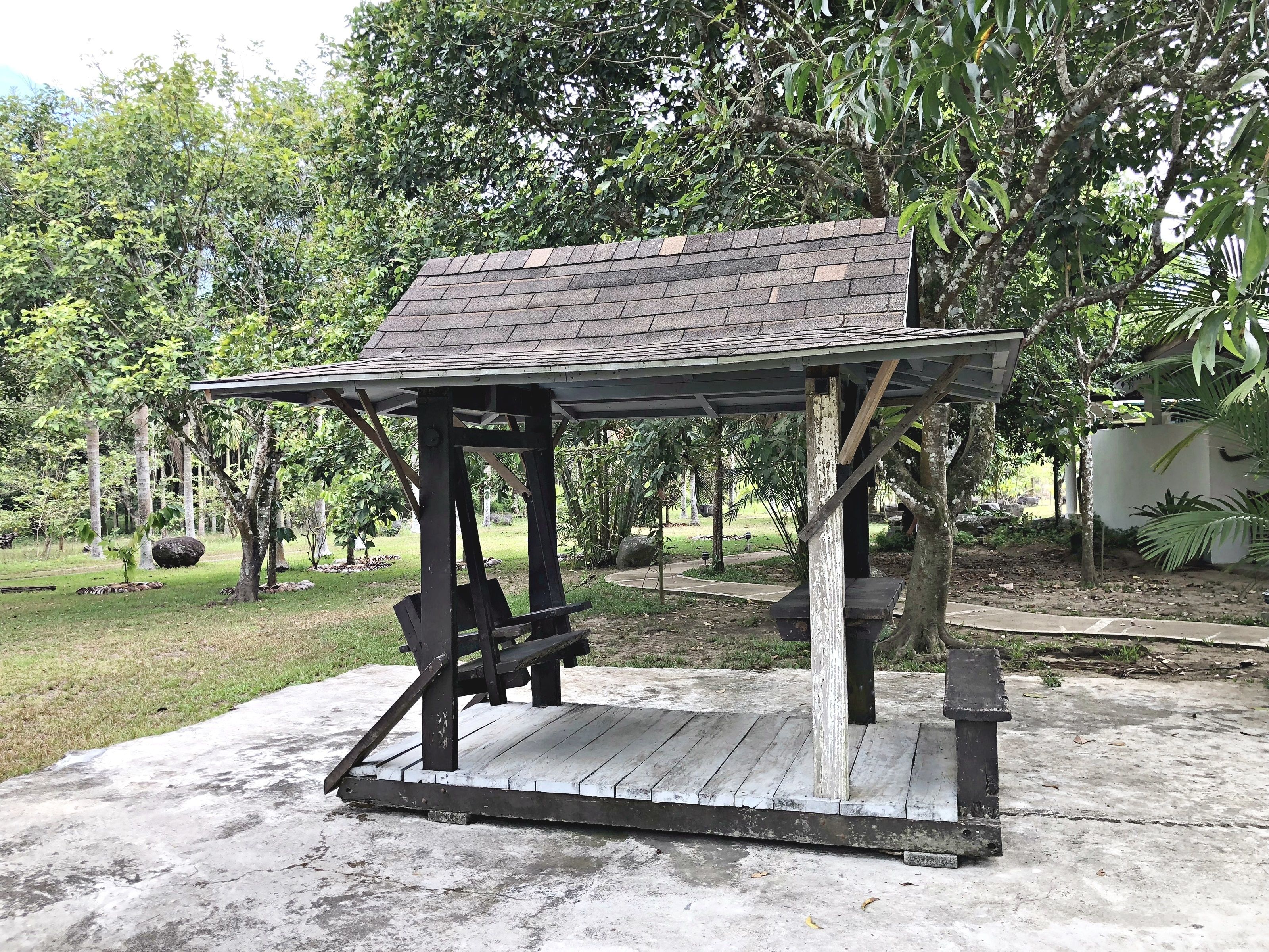 A wooden gazebo with a sloped roof, surrounded by trees and a grassy area.