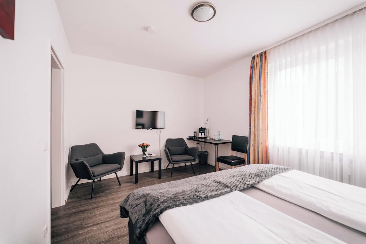Una camera d'albergo con un letto, due poltrone, un tavolino con un vaso, una scrivania con un computer e una televisione a parete.