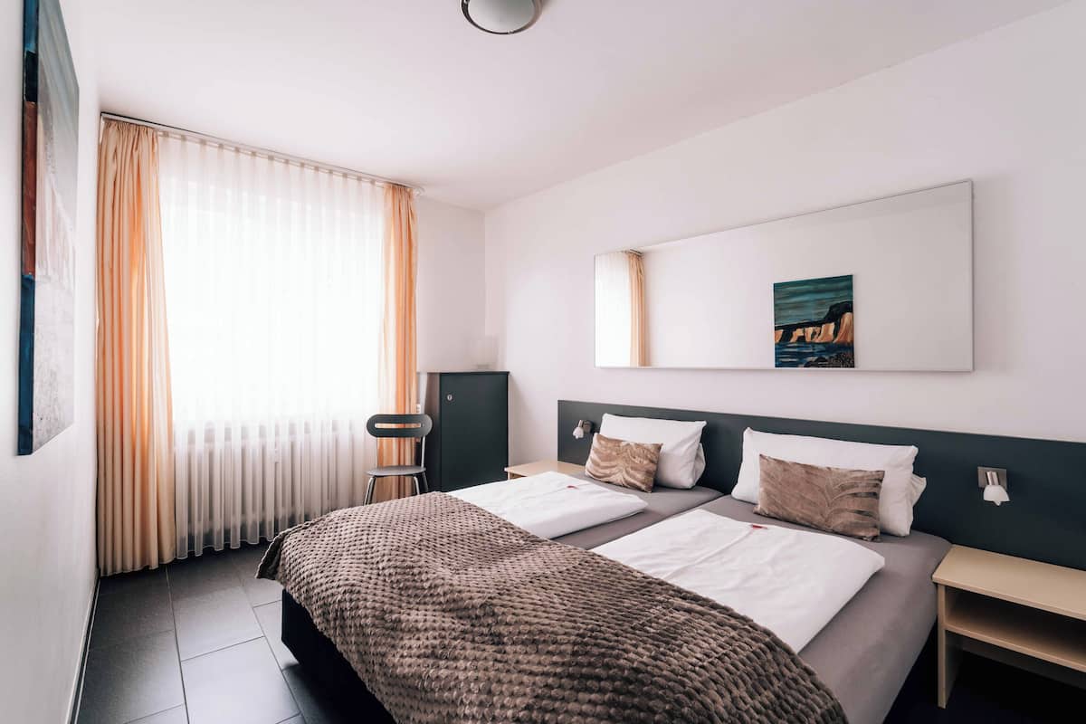 Una camera d'albergo con due letti, un'ampia finestra con tende, un quadro appeso al muro e un tavolino.