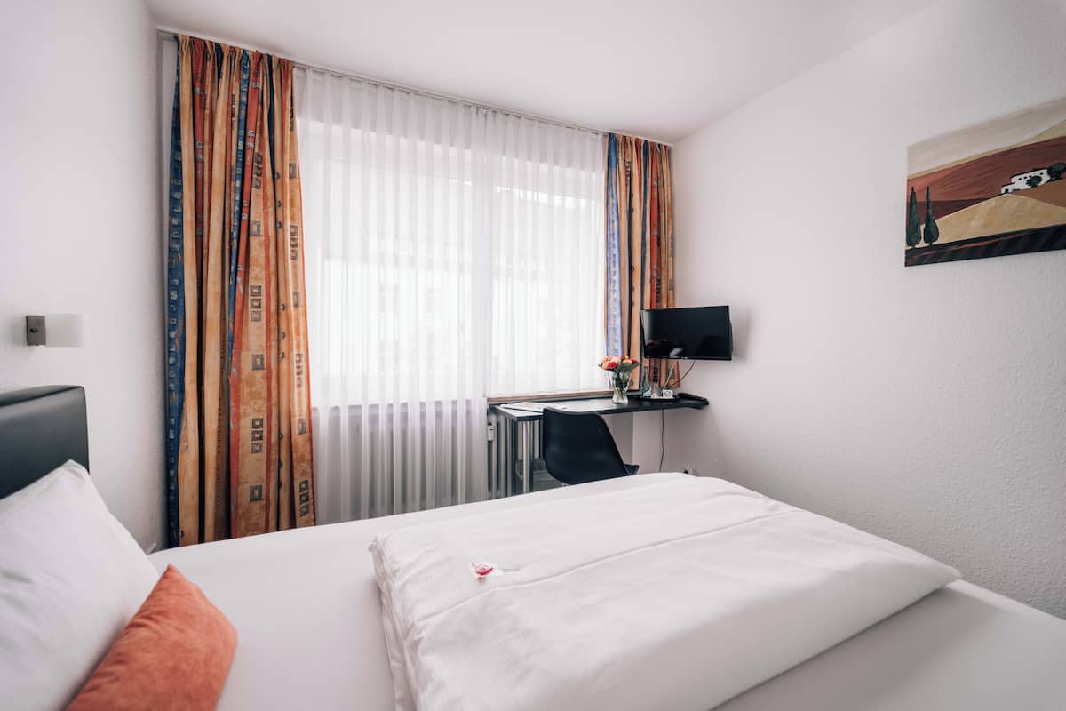 Una camera d'albergo con un letto, una scrivania, una sedia, una TV e una finestra con tende.
