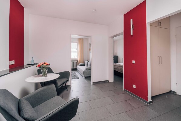 Una habitación de hotel moderna con un sofá gris, una mesita redonda con un florero y una pared de acento roja, además de vistas al área de estar contigua.