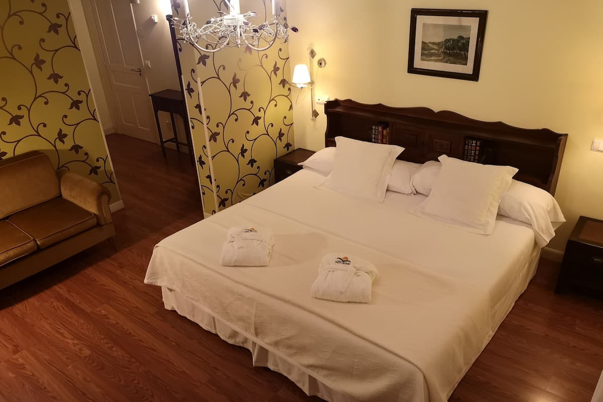 A neatly made bed with white linens and two towels, a wooden headboard, a chandelier, a framed picture on the wall, and a sofa in the corner.