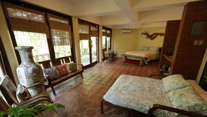 A spacious room with a large bed, a sofa, and a dining table. There are large windows with bamboo blinds, a decorative vase, and a potted plant.