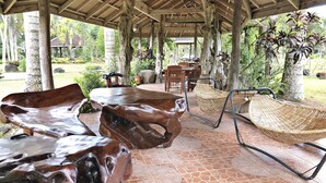 A covered outdoor area with a wooden table and chairs, a woven hammock, and a stone-tiled floor.