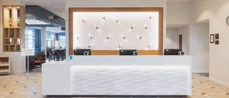 A hotel reception area with a modern design, featuring a large reception desk, a wall-mounted light fixture, and a visible sign for the bathroom.