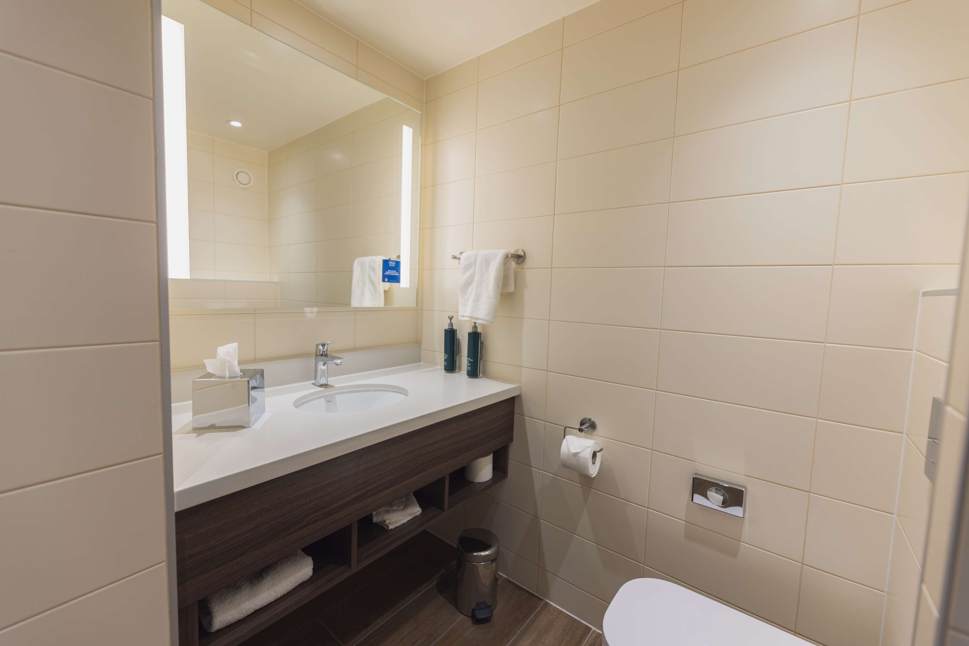 Un baño moderno con un espejo grande, un lavamanos y un inodoro. Hay toallas y artículos de aseo en la encimera.