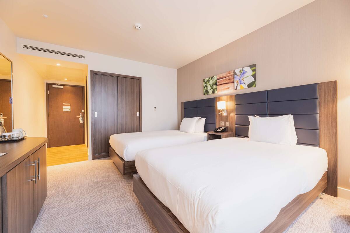 A hotel room with two beds, a wooden headboard, a bedside table with a lamp, and a door leading to a hallway.